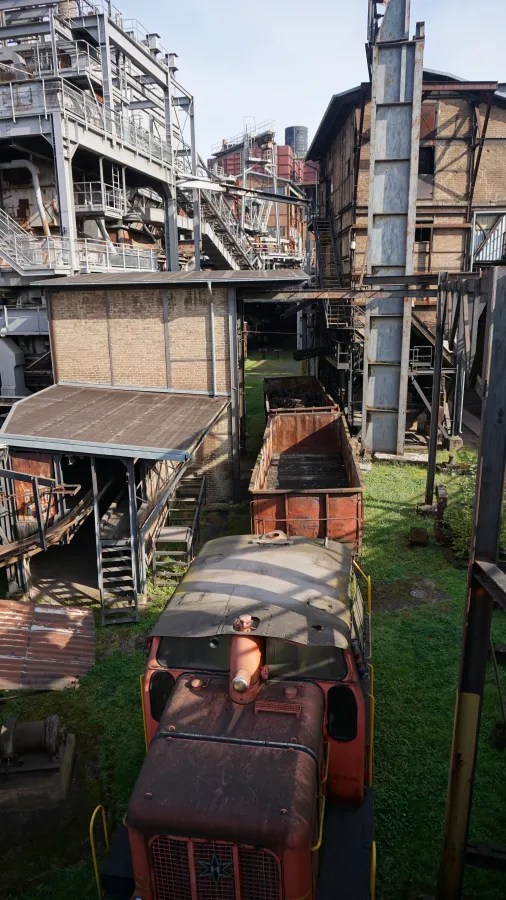 A view of industrial structures and a vintage train engine in an outdoor setting at Völklinger Hütte, highlighting the old railway carts and green grass.