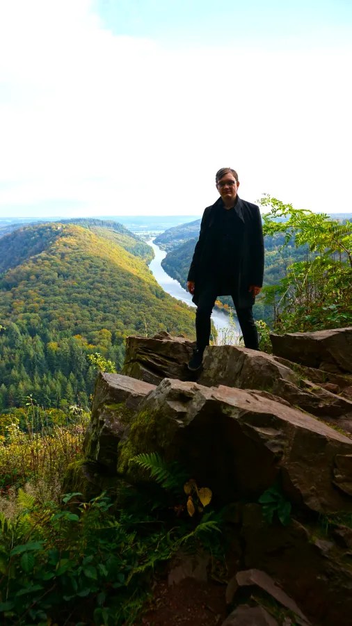 A person standing on a rocky ledge overlooking a lush green valley and river, with trees and hills in the background.