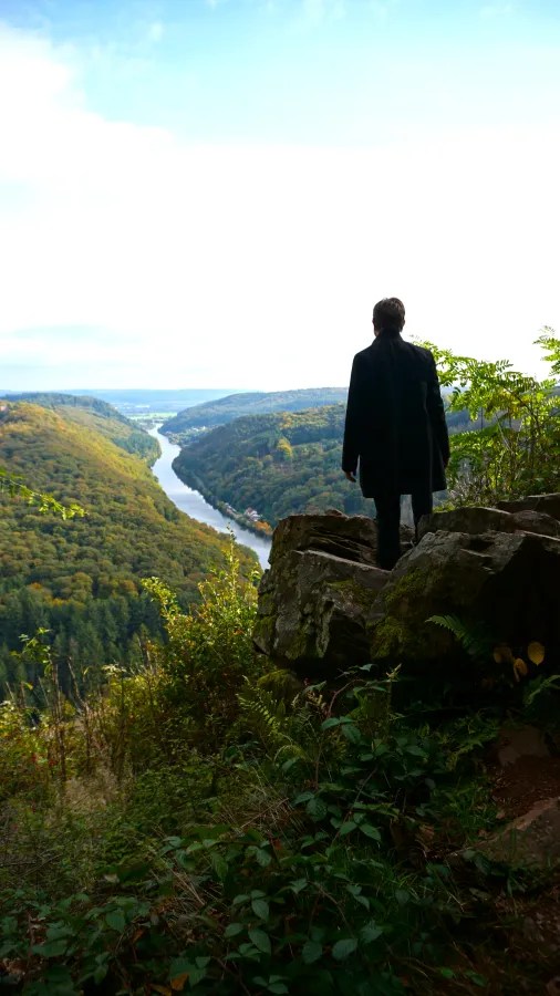 A person standing on a rocky outcrop overlooking a river winding through lush green hills under a cloudy sky.