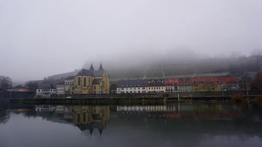 Eine malerische Stadt am Flussufer, umgeben von Nebel, mit historischen Gebäuden und einer Kirche im Vordergrund, die sich im Wasser spiegelt.