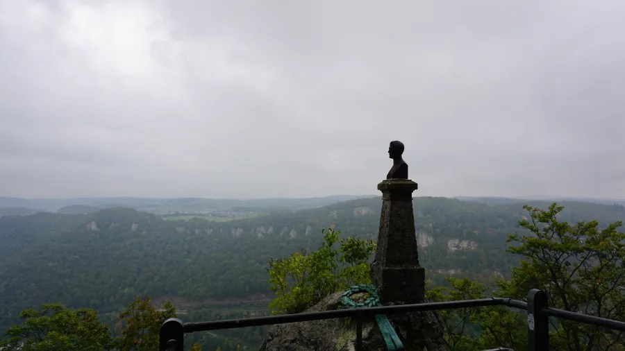 Eine Büste auf einem steineren Postament, die über eine bewaldete Landschaft mit einem nebligen Himmel hinausschaut.