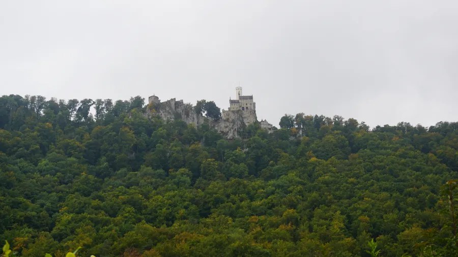 Ein Blick auf das Schloss Lichtenstein, das hoch oben auf einem Berg thront, umgeben von dichten Wäldern und unter einem bewölkten Himmel.