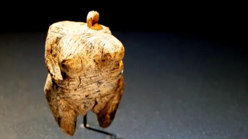 Eine kleine, geschnitzte Holzfigur, die eine menschliche Form darstellt, mit betonter Oberkörperstruktur und stehendem Kopf.