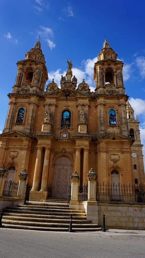 Fassade einer beeindruckenden Kirche in Malta mit zwei Türmen und kunstvollen Verzierungen unter einem blauen Himmel.
