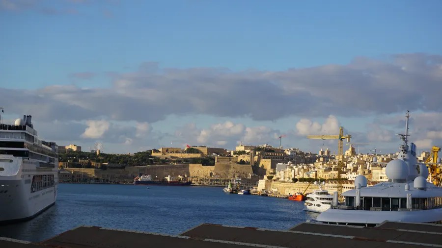 Blick auf den Hafen von Valletta mit Schiffen und der Altstadt im Hintergrund, unter blauem Himmel mit einigen Wolken.