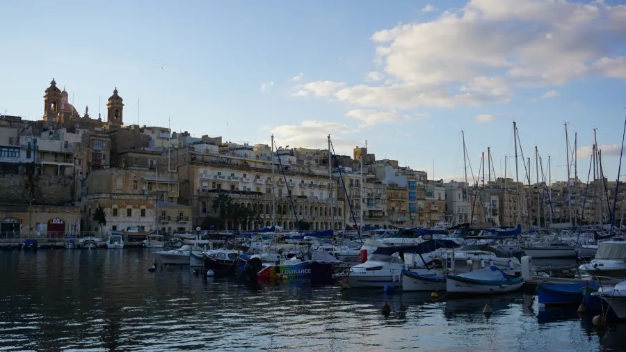 Blick auf den Hafen von Città Vittoriosa mit verschiedenen Booten, umgeben von historischen Gebäuden und einer bewölkten, aber klaren Himmel.