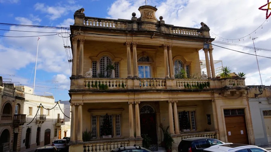 Eine alte, elegante Sandsteinfassade eines Hauses in Malta mit Säulen und einem geschmückten Balkon, umgeben von blauen Himmel und Wolken.