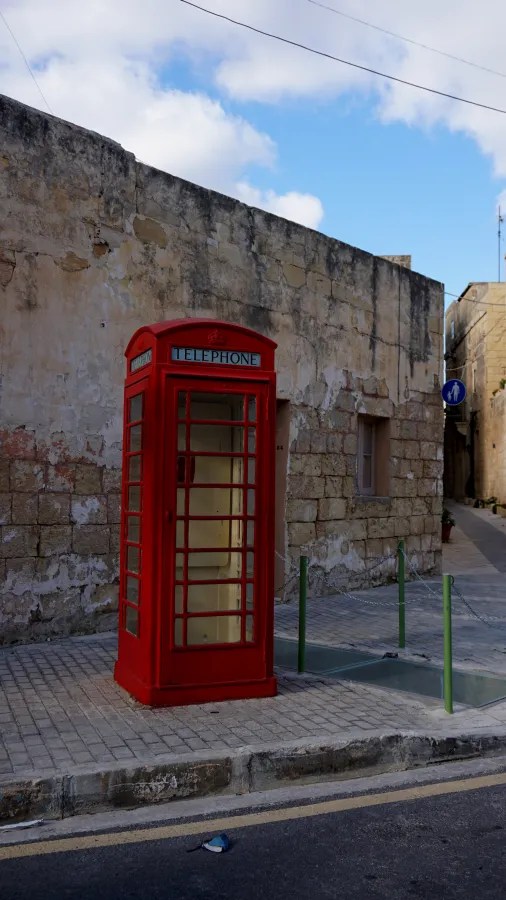 Rote Telefonzelle vor einer steinernen Wand in einer maltesischen Stadt, umgeben von blauen Himmel und schmalen Gassen.