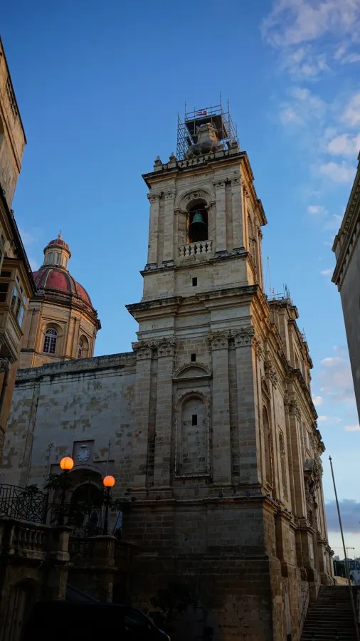 Eine historische Kirche mit einer hohen, verzierten Fassade und einem Glockenturm, umgeben von alten Gebäuden und schöner Himmel.