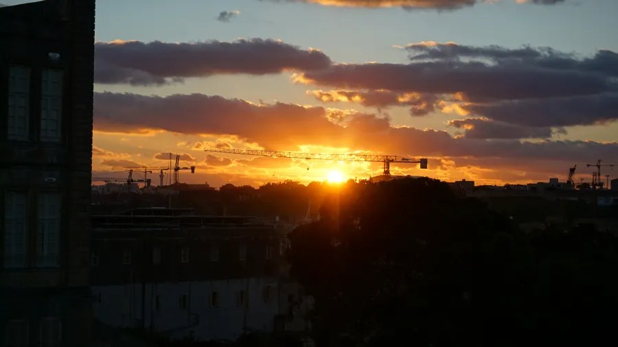 Sonnenuntergang hinter einer Stadtlandschaft mit Baukränen und Wolken am Himmel.