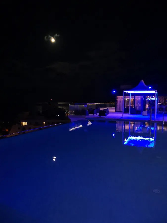 Eine nächtliche Aussicht auf einen beleuchteten Pool mit blauer Beleuchtung, einem Pavillon und dem Mond im Hintergrund.