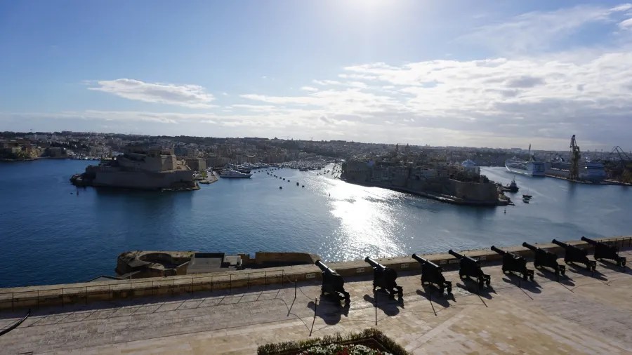 Blick auf den Hafen von Valletta mit historischen Kanonen im Vordergrund und der Stadt im Hintergrund, unter einem klaren blauen Himmel.