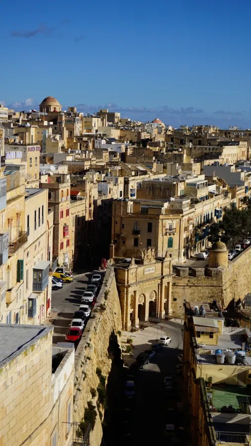 Panoramablick auf die Stadt Valletta mit ihren sandsteinfarbenen Gebäuden und engen Gassen, gesäumt von Autos auf der Straße, unter einem blauen Himmel.