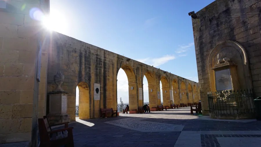 Blick auf eine alte Steinstruktur mit Bögen und Bänken, im Hintergrund strahlt die Sonne.