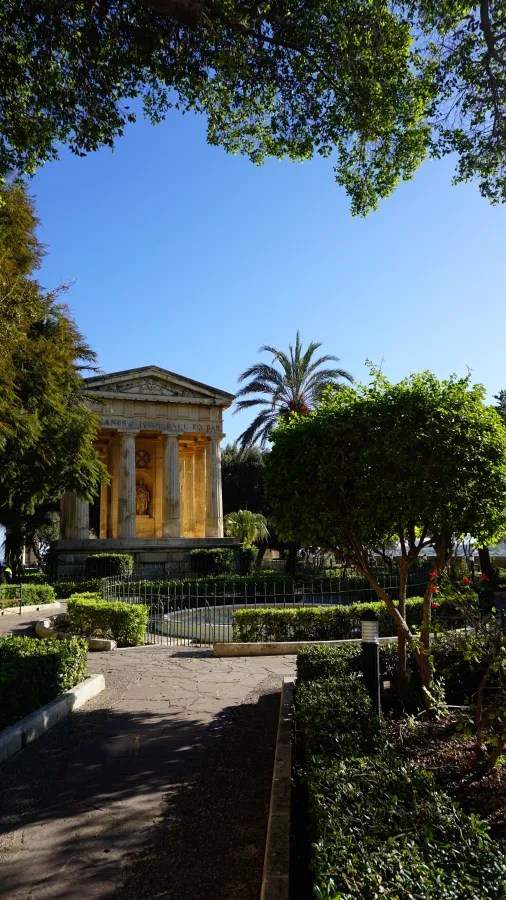Ein monumentales Gebäude mit Säulen im Zentrum eines gepflegten Gartens, umgeben von grünen Bäumen und Palmen, bei strahlend blauem Himmel.