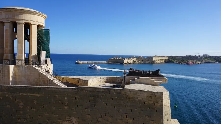 Blick auf das Wasser mit einem Boot, das an der Küste von Malta vorbeifährt, und einer historischen Architektur im Vordergrund.