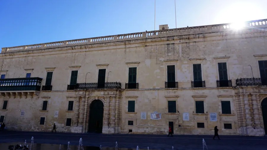 Historisches Gebäude in Valletta, Malta, mit lichtbeiger Fassade, grünen Fenstern und Balkon. Die Sonne scheint im Hintergrund.