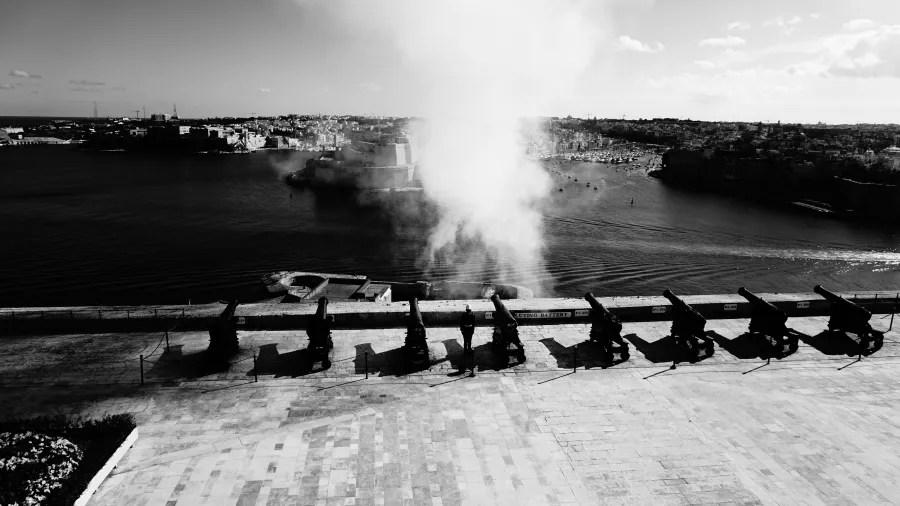 Blick auf den Hafen von Valletta mit Kanonen im Vordergrund und Rauch, der von einem abgefeuerten Kanonenschuss aufsteigt.