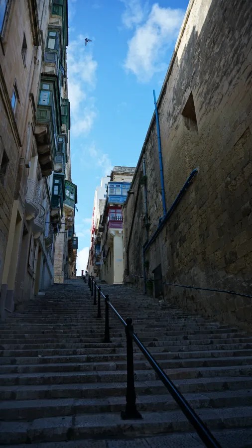 Blick auf eine steile Treppe, die von einer engen Gasse umgeben ist, mit historischen Gebäuden und blauen Balkonen auf beiden Seiten, unter einem strahlend blauen Himmel.