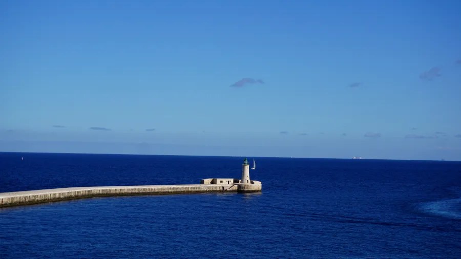Eine Aussicht auf das weitreichende blaue Meer mit einem kleinen Leuchtturm auf einer Landzunge im Vordergrund. Der Himmel ist klar und blau, mit wenigen Wolken.