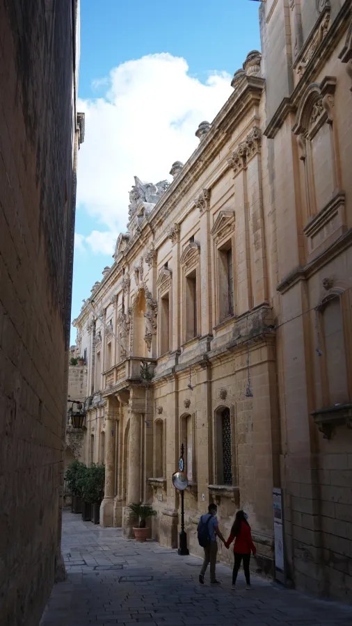 Ein romantisches Paar spaziert durch eine enge Gasse mit historischen Sandstein-Gebäuden in Malta unter einem blauen Himmel.