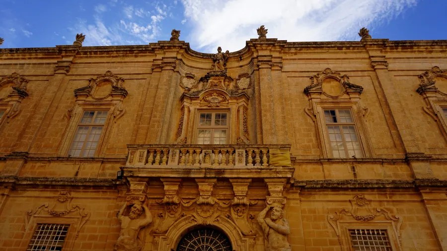 Historisches Gebäude in Malta mit dekorativer Fassade, Fenstern und Skulpturen im Stil der Barockarchitektur gegen einen blauen Himmel.