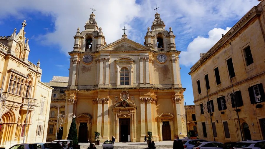 Ansicht der beeindruckenden Kirche mit zwei Türmen und kunstvollen Details, umgeben von historischen Gebäuden und einem klaren blauen Himmel in Malta.