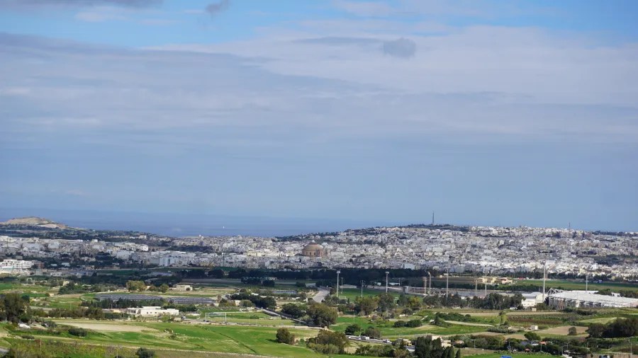 Panoramablick auf die Stadt mit weißen Gebäuden und umliegender Landschaft unter einem bewölkten Himmel.