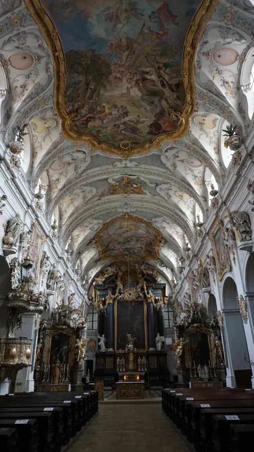 Innenansicht eines barocken Klosters mit dekorativem Deckenfresko, kunstvollen Statuen und einem zentralen Altar.