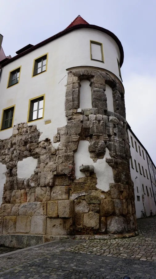 Detailansicht einer historischen Mauer mit einem modernen Gebäude in Regensburg, Deutschland. Die Mauer zeigt sichtbare Schäden und ist teilweise aus großen Steinen gebaut.