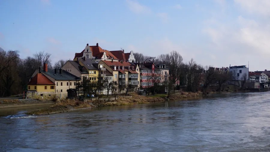 Blick auf Häuser am Ufer eines Flusses, mit Bäumen im Hintergrund und einem bewölkten Himmel.