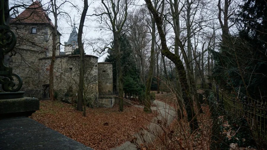 Eine schattige Wegführung durch einen herbstlichen, bewaldeten Bereich, mit steinernen Mauern und Türmen im Hintergrund.