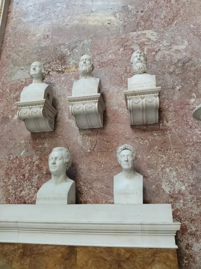 Mehrere weiße Büsten berühmter Persönlichkeiten auf einem marmorierten Hintergrund.