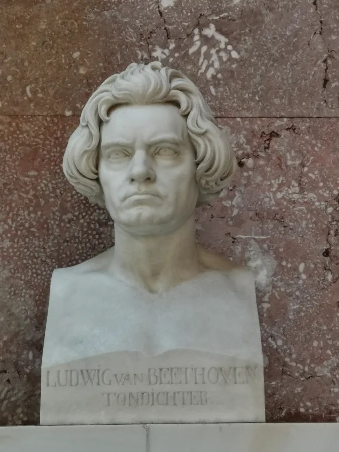 Eine weiße Marmorbüste von Ludwig van Beethoven, die auf einem Sockel steht, mit dem Schriftzug "LUDWIG VAN BEETHOVEN TONDICHTER" darunter. Im Hintergrund ist eine strukturierte, rötliche Wand zu sehen.