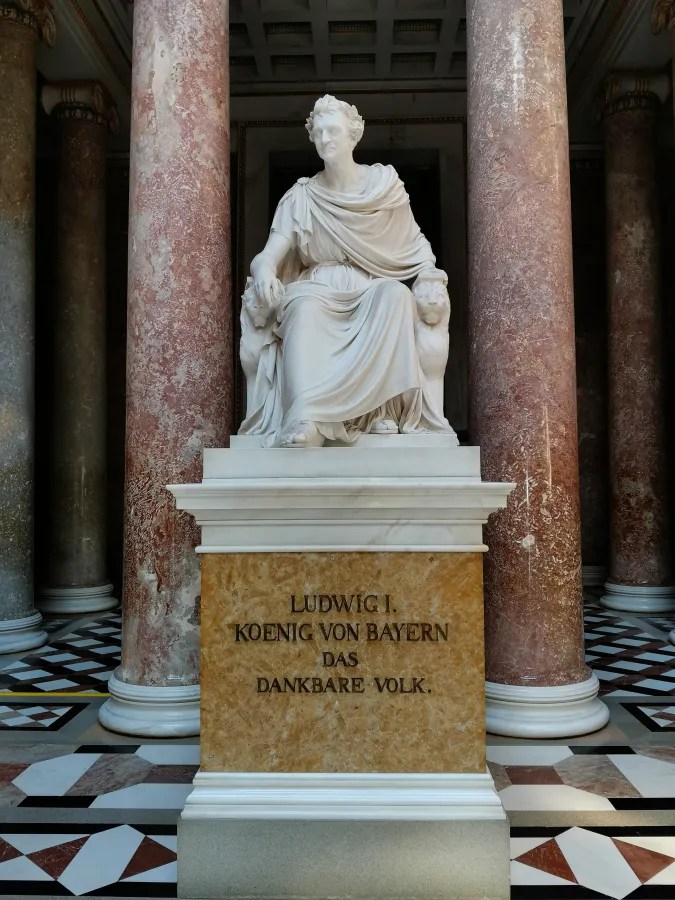 Eine Statue von Ludwig I., König von Bayern, die in einem beeindruckenden Innenraum sitzt, umgeben von Säulen und einem dekorativen Boden. Die Inschrift auf dem Sockel lautet: 'LUDWIG I. KOENIG VON BAYERN DAS DANKBARE VOLK.'
