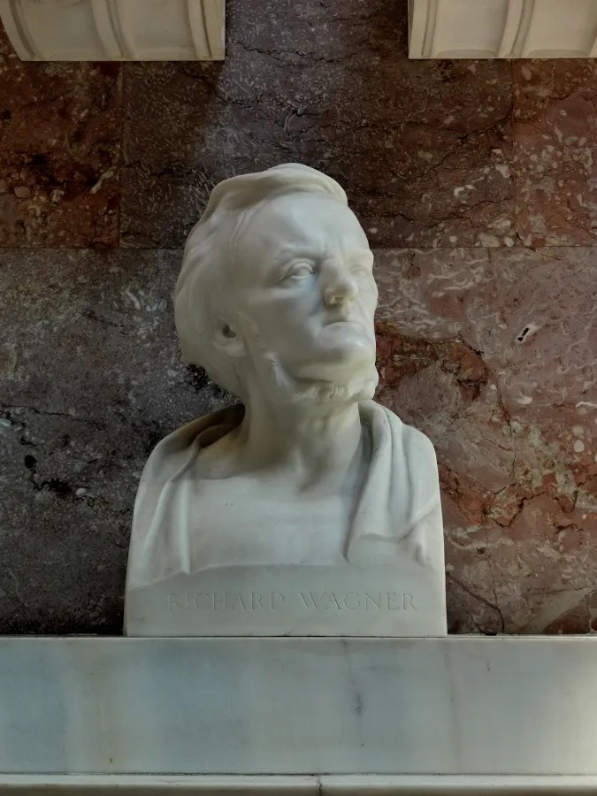 Eine Marmorbüste von Richard Wagner, die einen nachdenklichen Ausdruck zeigt, vor einem marmorierten Hintergrund.