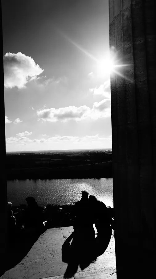 Silhouetten von Personen am Rand eines Gebäudes, das auf einen glitzernden Fluss und den Himmel mit Wolken blickt, während die Sonne hell scheint.