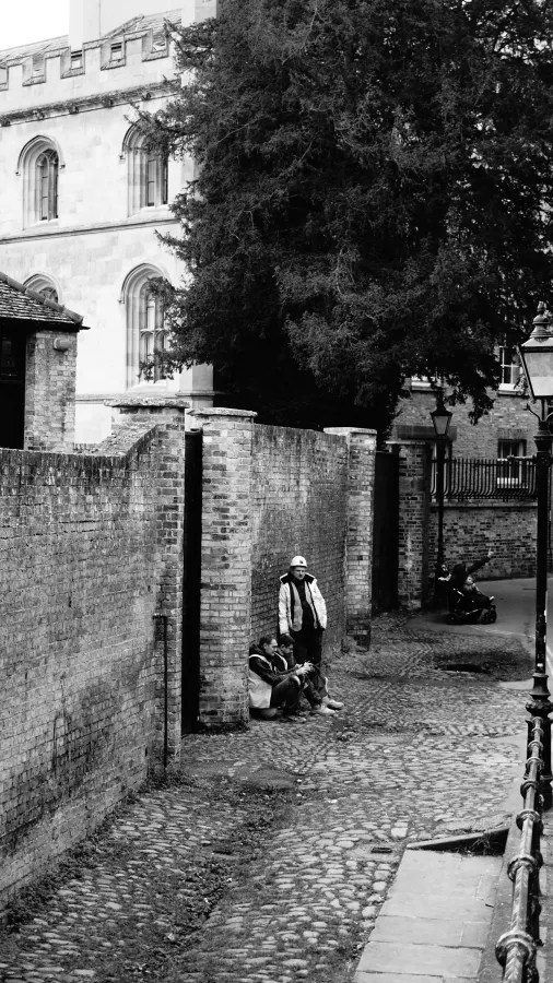 Schwarz-Weiß-Bild einer ruhigen Straße in Cambridge mit alten Ziegelmauern, einer Gruppe von Personen, die an einer Wand sitzen, und einem Mann in Arbeitskleidung, der daneben steht.