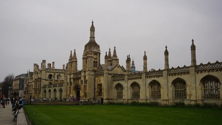 Blick auf die historische Architektur einer Universität in Cambridge mit neugotischen Elementen, umgeben von einer grünen Rasenfläche und einer grauen Himmelstruktur.