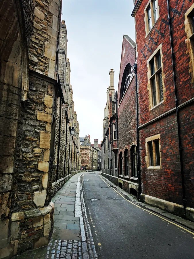 Schmale Gasse in Cambridge mit alten Steinhäusern und roten Backsteinfassaden, unter grauem Himmel.