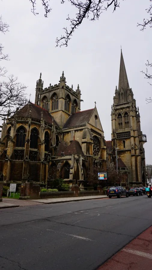 Ein Blick auf eine beeindruckende anglikanische Kirche mit gotischen Elementen in Cambridge. Die Kirche hat hohe Türme und verzierte Fassaden, umgeben von kahlen Bäumen und einer ruhigen Straße.