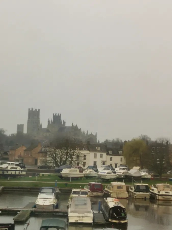 Blick auf eine bewölkte Hafenlandschaft in Cambridge mit Booten im Wasser und historischen Gebäuden im Hintergrund.