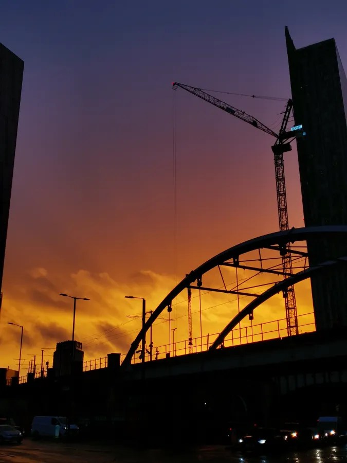 Sonnenuntergang hinter einer Stadt, sichtbar ist ein Baukran und eine freitragende Brücke, die vor einem orange-naturfarbenen Himmel silhouettiert sind.