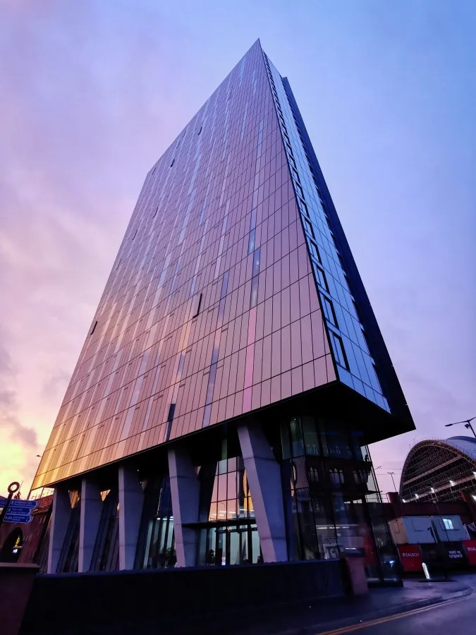 Moderne Glasfassade eines hohen Gebäudes bei Sonnenuntergang, mit reflektierenden Farben im Himmel.