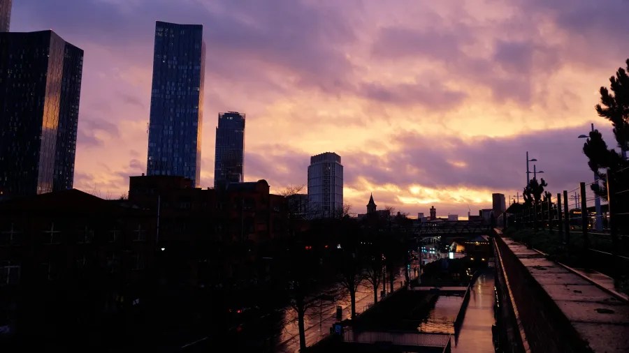 Silhouette der Skyline von Manchester bei Sonnenuntergang, mit modernen Hochhäusern im Vordergrund und dramatischen Wolken im Abendlicht.