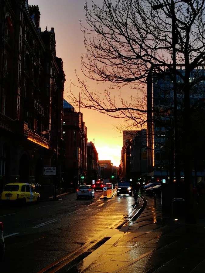 Sonnenuntergang über einer Stadtstraße in Manchester, reflektierende Pfützen auf dem Asphalt, moderne und historische Gebäude im Hintergrund, kahle Äste eines Baumes in der Silhouette.