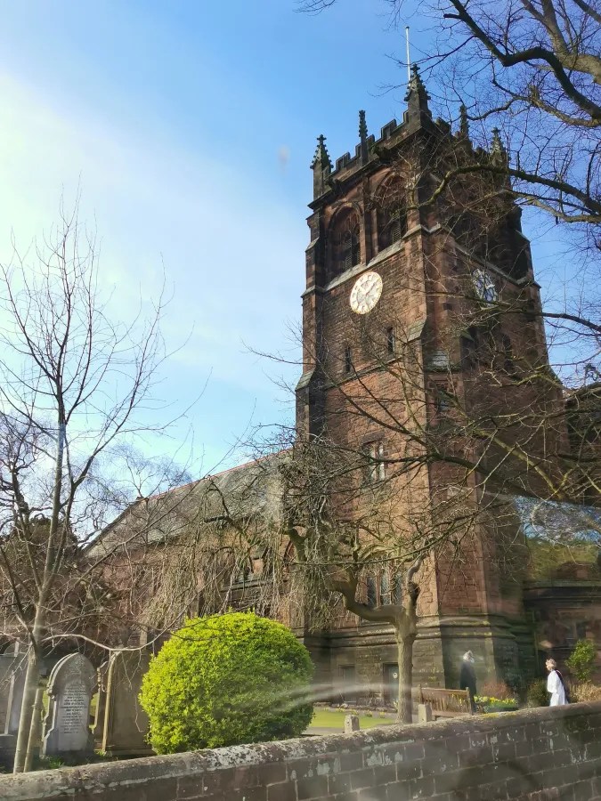 Eine alte Kirche mit einem markanten Turm und einer großen Uhr, umgeben von kahlen Bäumen und einem gepflegten Garten.