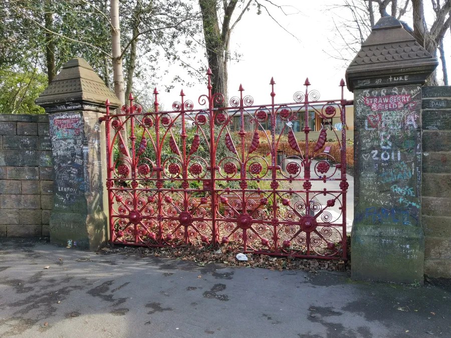 Ein rotes geschmiedetes Tor, das den Zugang zu einem Garten oder Park mit mehreren Bäumen und einer gepflasterten Fläche versperrt. Neben dem Tor sind Graffiti und Inschriften auf den Mauern sichtbar.