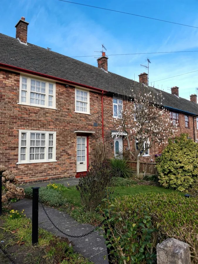 Ansicht eines Reihenhauses mit Ziegelsteinfassade, einem kleinen Vorgarten mit Pflanzen und einem Baum, unter klarem Himmel.