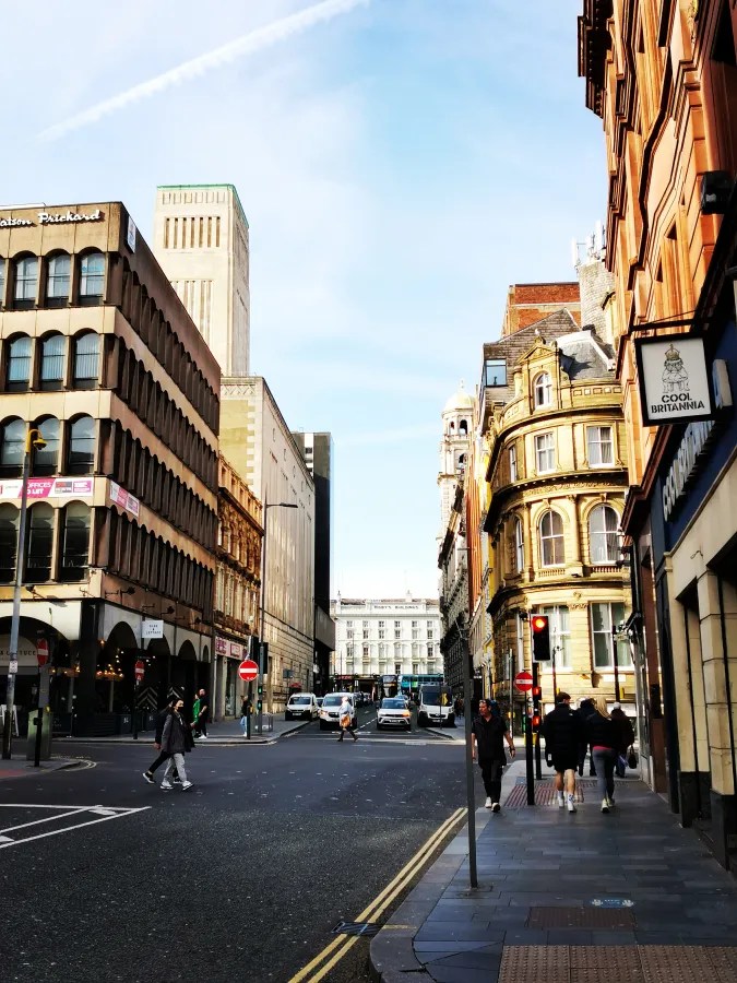 Stadtansicht mit einer belebten Straße, historischen und modernen Gebäuden, Verkehrsschildern und Fußgängern in einer urbanen Umgebung.